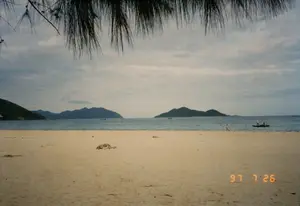Bức ảnh ghi lại một khung cảnh yên bình tại một bãi biển. Bãi cát vàng trải dài, phía trước là mặt biển xanh và ở phía xa là những hòn đảo hoặc dãy núi. Bầu trời có nhiều mây, cho thấy thời tiết không nắng gắt. Ở phía trên của bức ảnh, những cành lá của cây phi lao rủ xuống, tạo thành một cái khung tự nhiên cho phong cảnh. Trên mặt biển, có một chiếc thuyền nhỏ (ghe) với hai người đang ở trên đó, gần bờ. Có thể thấy một vài chiếc thuyền khác ở xa hơn. Trên bãi biển, một người, có vẻ là một đứa trẻ, đang đi bộ hoặc chạy gần mép nước. Ở góc dưới bên phải của bức ảnh, có dấu ngày tháng màu cam ghi '97 7 26', cho biết bức ảnh được chụp vào ngày 26 tháng 7 năm 1997.。（日本語訳）。写真は、あるビーチの穏やかな風景を記録しています。黄色い砂浜が広がり、その前には青い海面が、そして遠くには島々または山脈があります。空は雲が多く、天気が酷暑ではないことを示しています。写真の上部では、モクマオウの木の葉のついた枝が垂れ下がり、風景の自然なフレームを形成しています。海面には、岸の近くに2人が乗った小さなボート（ghe）が1隻あります。さらに遠くに他のボートが数隻見えます。ビーチでは、子供と思われる一人の人物が、水際近くを歩いているか走っています。写真の右下隅には、「97 7 26」と記されたオレンジ色の日付印があり、写真が1997年7月26日に撮影されたことを示しています。