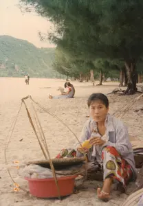 Bối cảnh bức ảnh là một bãi biển ở Việt Nam, có thể là khu vực gần Phan Rang, vào khoảng năm 1997. Bãi biển có dải cát rộng, phía sau là hàng cây phi lao và một ngọn đồi xanh ở đằng xa. Bầu trời có vẻ u ám.。Nhân vật chính ở tiền cảnh là một người phụ nữ bán hàng rong đang ngồi nghỉ trên cát. Cô đang ăn một thứ gì đó màu vàng, có thể là một miếng dứa (thơm) hoặc một trái bắp (ngô) luộc. Cô mặc trang phục giản dị với áo sơ mi khoác ngoài và quần hoa. Bên cạnh cô là bộ quang gánh, dụng cụ mưu sinh quen thuộc. Một bên quang gánh là một chiếc thau nhựa màu đỏ, bên trong đựng các món hàng được bọc trong túi ni lông, có khả năng là đồ ăn vặt hoặc hải sản để bán cho khách đi biển.。Ở hậu cảnh, có một vài người đang tận hưởng không khí biển. Một cặp đôi đang ngồi trên cát, và hai người khác đang đi dạo ở phía xa. Một chiếc xe đạp cũng có thể được nhìn thấy dựng gần hàng cây. Không có văn bản, biển báo hay chữ viết nào có thể nhận dạng trong ảnh.。（日本語訳）。写真の背景はベトナムのあるビーチで、おそらく1997年頃のファンラン近郊の地域です。ビーチには広い砂浜が広がり、その後ろにはモクマオウの並木と、遠くには緑の丘があります。空は曇っているようです。前景の主役は、砂浜で座って休んでいる一人の露天商の女性です。彼女は何か黄色いものを食べており、それはパイナップル（thơm）の一切れか、茹でたトウモロコシ（ngô）かもしれません。彼女はシャツを羽織り、花柄のズボンを履いた簡素な服装をしています。彼女の隣には、おなじみの生計を立てる道具である天秤棒があります。天秤棒の片方には赤いプラスチック製のたらいがあり、その中にはビニール袋に包まれた商品が入っています。おそらく、海水浴客に売るためのお菓子や海産物でしょう。後景には、数人の人々が海の雰囲気を楽しんでいます。一組のカップルが砂浜に座っており、他の二人が遠くを散歩しています。木々の近くに立てかけられた一台の自転車も見えます。写真の中には、識別できるテキスト、看板、または文字はありません。