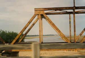 Bức ảnh được chụp từ một phương tiện đang di chuyển khi đi qua một cây cầu giàn thép. Cây cầu có kết cấu bằng kim loại, đã cũ và bị rỉ sét nhiều. Nhìn qua khung giàn của cây cầu là một vùng sông nước rộng lớn, có thể là một con sông hoặc cửa sông, với mực nước đang cạn, để lộ ra những bãi cát lớn. Ở phía xa, bên kia bờ là một dải đất với cây cối xanh tươi. Bầu trời có mây. Ở góc dưới bên phải của bức ảnh có dấu ngày tháng kỹ thuật số màu cam ghi '97 7 26', cho biết thời điểm chụp là ngày 26 tháng 7 năm 1997.。（日本語訳）。写真は、鋼鉄のトラス橋を渡る際に、移動中の乗り物から撮影されたものです。その橋は金属構造で、古く、ひどく錆びています。橋のトラス構造のフレーム越しには、広大な水域が見えます。それは川か河口かもしれず、水位は低く、大きな砂州が露出しています。遠くの対岸には、青々とした木々が生い茂る一帯の土地があります。空は曇っています。写真の右下隅には、「'97 7 26」と記されたオレンジ色のデジタル日付スタンプがあり、撮影日時が1997年7月26日であることを示しています。