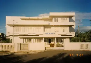 Bức ảnh chụp một tòa nhà lớn màu trắng vào một ngày nắng đẹp, ngày 27 tháng 7 năm 1997. Đây là một Nhà khách (Guest House), có thể là một cơ sở lưu trú của nhà nước. Trên mặt tiền tòa nhà có một biển hiệu màu xanh dương ghi rõ chữ 'NHÀ KHÁCH' và bên dưới là 'GUEST HOUSE'. Phía trên có một dòng chữ nhỏ hơn nhưng không thể đọc được. Tòa nhà có kiến trúc hiện đại, nhiều tầng với nhiều cửa sổ có song sắt. Toàn bộ công trình được bao quanh bởi một hàng rào bê tông với các khối thông gió trang trí và một chiếc cổng sắt đã hơi gỉ. Phía trước là một con đường trải nhựa. Hai bên tòa nhà có hai cột đèn đường cao. Phía sau bên phải có thể thấy những hàng cây dừa. Bầu trời trong xanh, quang đãng. Khung cảnh tĩnh lặng, không có hoạt động của con người.。（日本語訳）。1997年7月27日の晴れた日に撮影された、大きな白い建物の写真。これは迎賓館（ゲストハウス）であり、国営の宿泊施設である可能性がある。建物の正面には「NHÀ KHÁCH」と明記され、その下に「GUEST HOUSE」と書かれた青い看板がある。その上にはもっと小さな文字の列があるが、読むことはできない。建物は近代的な建築様式で、多くの階があり、鉄格子のついた窓がたくさんある。建物全体は、装飾的な通気ブロックのあるコンクリートの塀と、少し錆びた鉄の門で囲まれている。正面にはアスファルトの道路がある。建物の両側には、2本の高い街灯がある。空は青く澄み渡っている。風景は静かで、人の活動は見られない。