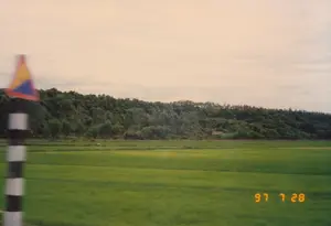 Bức ảnh chụp một phong cảnh nông thôn ở Việt Nam, có thể là ở khu vực Quảng Bình hoặc Quảng Trị, vào ngày 28 tháng 7 năm 1997. Cảnh vật chính là một cánh đồng lúa xanh mướt trải dài. Ở phía sau là một ngọn đồi được bao phủ bởi cây cối rậm rạp. Thấp thoáng có một vài ngôi nhà mái ngói nằm dưới chân đồi. Ở bên trái bức ảnh có một cột mốc giao thông sơn sọc đen trắng, phía trên là một biển báo hình tam giác bị mờ do chuyển động nên không thể đọc được nội dung. Góc dưới bên phải có dấu ngày tháng màu vàng ghi '97 7 28'. Khung cảnh cho thấy bức ảnh được chụp từ một phương tiện đang di chuyển ven đường.。（日本語訳）。写真は1997年7月28日にベトナムの田舎の風景を撮影したもので、おそらくクアンビン省かクアンチ省の地域です。主な景色は、広がる青々とした水田です。後方には、木々が鬱蒼と茂る丘があります。丘の麓には、瓦屋根の家がいくつか見え隠れしています。写真の左側には、白黒の縞模様に塗られた交通標識の柱があり、その上には、動きによってぼやけて内容が読み取れない三角形の標識があります。右下隅には、「'97 7 28」と記された黄色い日付スタンプがあります。この光景は、写真が道端を移動中の乗り物から撮影されたことを示しています。