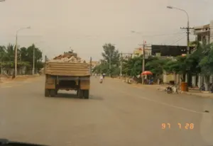 Bức ảnh chụp cảnh một con đường tại một thị trấn hoặc thành phố ở Việt Nam, được chụp từ bên trong một chiếc ô tô đang di chuyển vào ngày 28 tháng 7 năm 1997. Ở trung tâm bức ảnh là phía sau của một chiếc xe tải ben đang chạy trên đường, chở đầy vật liệu có thể là đá hoặc xà bần. Con đường được trải nhựa, hai bên đường là vỉa hè đất, có trồng cây xanh, cột điện và đèn đường. Dọc theo con đường là những ngôi nhà và cửa hàng. Phía bên phải, có thể thấy một quán nước nhỏ ven đường với chiếc ô màu đỏ, một vài người đang ngồi, một chiếc xe máy đang đậu và một thùng rác công cộng màu cam. Ở phía xa, có những người khác đang đi xe máy. Có một số biển hiệu gắn trên cột điện nhưng chữ viết không thể đọc được. Dấu ngày tháng '97 7 28' được hiển thị ở góc dưới bên phải của hình ảnh.。（日本語訳）。1997年7月28日に、走行中の車内から撮影された、ベトナムのある町または都市の道路の光景を写した写真です。写真の中央には、道路を走行しているダンプカーの後ろ姿があり、石か建設廃材と思われる資材を満載しています。道路は舗装されており、両側は土の歩道で、街路樹、電柱、街灯があります。道に沿って家や店が並んでいます。右側には、赤いパラソルのある道端の小さな飲み物屋、座っている数人の人々、停まっている一台のバイク、そしてオレンジ色の公共ゴミ箱が見えます。遠くには、バイクに乗っている他の人々がいます。電柱にはいくつかの看板が取り付けられていますが、文字は読み取れません。画像の右下隅に「97 7 28」という日付スタンプが表示されています。