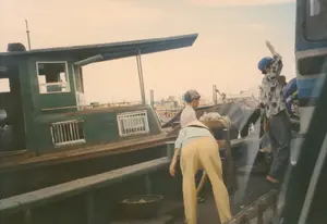 Bối cảnh trong ảnh là một bến cảng hoặc bến sông, có nhiều tàu thuyền đang neo đậu. Nổi bật là một chiếc tàu lớn sơn màu xanh lá cây với cabin. Có nhiều người trong ảnh, phản ánh một cảnh sinh hoạt đời thường. Một người phụ nữ đội nón lá, mặc áo màu xanh nhạt và quần vàng, đang cúi người, có thể là đang chuyển hàng hoặc lên xuống thuyền. Gần đó có hai cậu bé. Một cậu bé đội mũ lưỡi trai đang đứng nhìn. Một cậu bé khác đội mũ bảo hiểm màu xanh, đứng trên thành của một phương tiện (có thể là xe tải hoặc xe khách), đang giơ tay lên như đang vẫy chào. Có một chiếc giỏ đan được đặt gần người phụ nữ. Không có chữ viết nào có thể nhận dạng được trong ảnh.。（日本語訳）。写真の背景は港または川岸で、多くの船が停泊しています。目立っているのは、船室のある緑色に塗られた大きな船です。写真には多くの人が写っており、日常の生活風景を反映しています。ノンラーをかぶり、水色の服と黄色いズボンを履いた女性が、荷物を運んでいるか、船に乗り降りしているのか、身をかがめています。その近くに二人の少年がいます。キャップをかぶった少年が立って見ています。もう一人の少年は青いヘルメットをかぶり、乗り物（トラックかバスの可能性があります）の縁に立って、挨拶するかのように手を挙げています。女性の近くに編みかごが置かれています。写真には識別できる文字はありません。