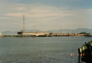 Trên ảnh có dấu ngày "97 7 29", cho thấy ảnh được chụp vào ngày 29 tháng 7 năm 1997. Khung cảnh chính là một công trường xây dựng một cây cầu lớn bằng bê tông đang bắc qua một con sông hoặc cửa sông rộng. Cây cầu đang trong quá trình thi công, với nhiều cần cẩu và thiết bị xây dựng trên mặt cầu và trên các sà lan neo đậu gần các trụ cầu. Xa xa phía sau cây cầu là dãy núi. Bên bờ sông có thể thấy một cột điện cao thế và cây cối. Trên mặt nước có một vài chiếc thuyền nhỏ đang hoạt động. Ở phía trước, góc dưới bên phải, là một phần của một con tàu lớn.。（日本語訳）。写真には「97 7 29」という日付スタンプがあり、写真が1997年7月29日に撮影されたことを示している。主な風景は、広い川または河口に架かる大きなコンクリート橋の建設現場である。橋は建設中であり、橋の上や橋脚の近くに停泊しているバージ船の上には、多くのクレーンや建設機械がある。遠く、橋の後ろには山脈がある。水面では数隻の小さな船が活動している。手前の右下隅には、大きな船の一部がある。