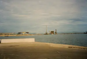 Bức ảnh chụp một công trường xây dựng quy mô lớn, dường như là một cây cầu đang được xây dựng bắc qua một vùng nước rộng như vịnh, cửa sông hoặc biển. Ở phía trước là một bờ kè hoặc bến tàu bằng bê tông, trên đó có một sợi dây xích sắt. Trên mặt nước, có nhiều kết cấu xây dựng lớn đang trong quá trình thi công, bao gồm các trụ cầu, giàn giáo phức tạp, và các sà lan công trình. Có thể thấy các cần cẩu và máy móc hạng nặng đang hoạt động. Ở phía bên trái, một đoạn dầm cầu dài đang được lắp đặt. Một chiếc thuyền nhỏ (ghe) đang di chuyển trên mặt nước gần bờ. Phía xa là đường bờ biển với những dãy núi mờ ảo. Bầu trời u ám, có nhiều mây. Không có văn bản nào có thể đọc được trong ảnh.。（日本語訳）。写真は大規模な建設現場を撮影したもので、湾、河口、または海のような広い水域を渡る橋が建設されているようです。手前にはコンクリート製の護岸または埠頭があり、その上には鉄の鎖があります。水上では、橋脚、複雑な足場、作業用の台船を含む、多くの大規模な建設構造物が工事中です。クレーンや重機が稼働しているのが見えます。左側では、長い橋桁の一部が設置されています。小さなボート（ゲ）が岸近くの水面を移動しています。遠くには、ぼんやりとした山々のある海岸線が見えます。空は曇っており、雲が多いです。写真には読み取れる文字はありません。