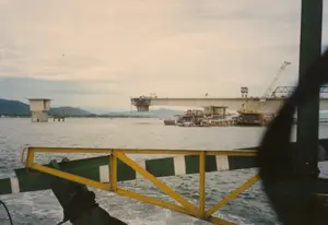 Bức ảnh ghi lại cảnh một công trình xây dựng cầu lớn bắc qua một con sông hoặc cửa sông rộng. Cây cầu đang trong quá trình thi công dở dang, với các trụ cầu bê tông và các nhịp cầu chưa được nối liền. Một trụ cầu đứng trơ trọi giữa dòng nước. Có thể thấy các cần cẩu và máy móc xây dựng đang hoạt động trên công trường. Dưới chân cầu, một số tàu thuyền, có thể là tàu công trình hoặc thuyền nhà, đang neo đậu. Phía xa là dãy núi và bờ sông. Bức ảnh được chụp từ trên một chiếc phà hoặc một con tàu đang di chuyển, có thể thấy qua phần lan can màu vàng và thành tàu màu xanh lá cây-trắng ở phía trước. Mặt nước xung quanh đang sủi bọt, cho thấy phương tiện này đang chạy.。（日本語訳）。写真は、広い川または河口に架かる大きな橋の建設現場の光景を記録しています。橋は建設途中の段階にあり、コンクリートの橋脚とまだ接続されていない橋桁があります。一つの橋脚が水流の真ん中にぽつんと立っています。建設現場ではクレーンや建設機械が稼働しているのが見えます。橋の麓では、作業船かハウスボートと思われるいくつかの船が停泊しています。遠くには山脈と川岸が見えます。写真は移動中のフェリーまたは船の上から撮影されたもので、手前にある黄色の手すりと緑と白の船体からそれがわかります。周囲の水面は泡立っており、この乗り物が航行中であることを示しています。