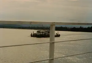 Bức ảnh chụp cảnh một chiếc phà đang vận chuyển hành khách và phương tiện qua một con sông rộng. Trên phà có ít nhất một chiếc xe khách lớn màu trắng, và có vẻ như có một chiếc nữa bên cạnh. Nhiều người đang đứng trên boong phà, xung quanh chiếc xe khách. Đây là một hình thức giao thông đường thủy phổ biến ở Việt Nam thời kỳ này, dùng để vượt sông ở những nơi chưa có cầu. Khung cảnh được chụp từ một vị trí cao, có thể là từ trên một cây cầu, với phần lan can màu trắng ở phía trước. Phía xa là bờ sông với cây cối rậm rạp và dãy núi mờ ảo dưới bầu trời u ám. Không có văn bản nào có thể đọc được trong ảnh.。（日本語訳）。写真は、一隻のフェリーが乗客と車両を乗せて広い川を渡っている光景を写したものです。フェリーの上には、少なくとも一台の大きな白いバスがあり、その隣にもう一台あるようです。多くの人々がバスの周りのフェリーの甲板に立っています。これは、この時代のベトナムで一般的な水上交通の一形態であり、まだ橋がない場所で川を渡るために使われます。この光景は高い場所から撮影されたもので、おそらく橋の上からで、手前には白い手すり部分があります。写真の中には読み取れる文字はありません。