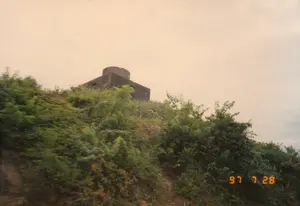 Trong ảnh là một công trình kiến trúc quân sự cũ, có thể là một lô cốt hoặc một đài quan sát, nằm trên đỉnh một ngọn đồi. Công trình được xây bằng đá, có phần thân hình khối và phần đỉnh hình tròn. Xung quanh là cây cối và thảm thực vật rậm rạp, bao phủ gần như toàn bộ sườn đồi. Bầu trời phía sau có màu vàng đục, có thể là do thời tiết hoặc do màu của tấm ảnh cũ. Ở góc dưới bên phải của ảnh có dấu ngày tháng màu cam "97 7 28". Khung cảnh cho thấy đây là một di tích lịch sử, có thể là từ thời chiến tranh, nằm giữa thiên nhiên hoang sơ.。（日本語訳）。写真には、丘の頂上にある古い軍事建築物が写っている。それはトーチカか監視塔かもしれない。その建造物は石で建てられており、胴体部分は角柱状で、頂上部分は円形である。周囲は木々や生い茂る植物で、丘の斜面全体をほぼ覆っている。後ろの空は濁った黄色をしており、天候のせいか、あるいは古い写真の色合いのせいかもしれない。写真の右下隅には、「97 7 28」というオレンジ色の年月日のスタンプがある。この光景は、これが手つかずの自然の中に位置する、おそらくは戦争時代からの歴史的遺物であることを示している。