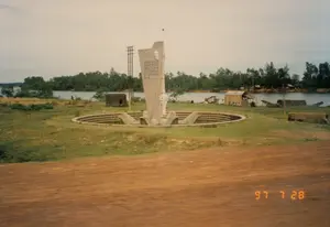 Bức ảnh chụp một khu đài tưởng niệm lịch sử nằm bên bờ sông vào ngày 28 tháng 7 năm 1997. Ở trung tâm là một tượng đài lớn bằng bê tông, bao gồm một tấm bia cao có khắc chữ, một bức phù điêu chân dung (dường như là Chủ tịch Hồ Chí Minh) và một ngôi sao năm cánh trên đỉnh. Phía trước tấm bia là bức tượng một người lính trong tư thế đứng gác, tay cầm súng. Toàn bộ cụm tượng đài được đặt trong một công trình hình tròn có các bậc thang đi xuống. Dòng chữ khắc trên bia đá, tuy hơi mờ, nhưng có thể nhận ra một phần nội dung là câu nói nổi tiếng: "NHIỆM VỤ CỦA THANH NIÊN KHÔNG PHẢI LÀ HỎI NƯỚC NHÀ ĐÃ CHO MÌNH NHỮNG GÌ, MÀ PHẢI TỰ HỎI MÌNH ĐÃ LÀM GÌ CHO NƯỚC NHÀ...". Phía sau đài tưởng niệm là một con sông rộng, bên kia bờ là cây cối um tùm. Dọc bờ sông phía bên phải có một vài ngôi nhà tạm, lán trại đơn sơ và một số chiếc thuyền (ghe) đang neo đậu. Gần tượng đài còn có một công trình nhỏ giống lô cốt và một cột điện. Tiền cảnh là một con đường đất bị nhòe, cho thấy ảnh được chụp từ một phương tiện đang di chuyển. Góc dưới bên phải có dấu ngày tháng màu cam "97 7 28".。（日本語訳）。1997年7月28日に川岸にある歴史記念碑地区を撮影した写真。中央にはコンクリート製の大きな記念碑があり、文字が刻まれた高い石碑、肖像のレリーフ（ホー・チ・ミン主席と思われる）、そして頂上には五芒星がある。石碑の前には、銃を手に警備の姿勢で立つ兵士の像がある。石碑に刻まれた文字は、少しぼやけているが、その内容の一部は有名な言葉であることがわかる：「青年の任務は、国が自分に何を与えてくれたかを問うことではなく、自分が国のために何をしたかを自問することである…」。右側の川岸沿いには、いくつかの仮設住宅、簡素な小屋、そして停泊している数隻の船（小舟）がある。記念碑の近くには、トーチカのような小さな建造物と電柱もある。前景にはぼやけた土の道があり、写真が移動中の乗り物から撮影されたことを示している。右下隅にはオレンジ色の日付スタンプ「97 7 28」がある。