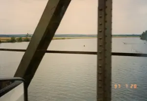 写真は、鉄骨構造の橋を渡っている時、移動中の乗り物、おそらく自動車から撮影されたものです。橋の梁の間から広大な川が見えます。川面には、赤いブイと仕掛けられた漁網が見え、地元の人々の水産漁業活動を示しています。遠くの川岸は、田畑、木々、そして遠くに電柱がある農村地帯です。写真の右下隅にはオレンジ色の「97 7 28」という日付スタンプがあり、撮影日時が1997年7月28日であることを示しています