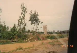 Bức ảnh chụp một khung cảnh ở vùng nông thôn, có thể là tại một di tích lịch sử, được chụp từ bên trong một phương tiện đang di chuyển. Ở góc dưới bên phải có dấu ngày tháng màu cam "97 7 28", cho biết ảnh được chụp vào ngày 28 tháng 7 năm 1997.。Ở trung tâm bức ảnh, phía xa là một tượng đài lớn bằng đá hoặc bê tông, có kiến trúc hiện đại. Trên thân tượng đài có khắc nhiều chữ nhưng không thể đọc rõ nội dung do khoảng cách và chất lượng ảnh. Dựa vào bối cảnh địa lý (Quảng Trị), đây có thể là một đài tưởng niệm chiến tranh, ví dụ như Tượng đài Khát vọng Thống nhất ở bờ Nam sông Bến Hải.。Phía sau bên phải tượng đài là một cây cầu. Phong cảnh xung quanh khá hoang sơ với con đường đất ở tiền cảnh. Cây cối bao gồm các cây cao như cây bạch đàn và một vài bụi chuối ở bên trái. Phía xa bên trái còn có một cột điện. Cạnh phải của ảnh bị che bởi khung cửa sổ xe, cho thấy người chụp đang ở trong ô tô hoặc xe buýt.。（日本語訳）。この写真は、田舎の風景、おそらく史跡で撮影されたもので、移動中の乗り物の中から撮られたものです。右下隅にはオレンジ色の「97 7 28」という日付印があり、写真が1997年7月28日に撮影されたことを示しています。写真の中央、遠方には、石またはコンクリートでできた、近代的な建築様式の大きな記念碑があります。記念碑の本体には多くの文字が刻まれていますが、距離と画質のため、内容ははっきりと読み取れません。地理的背景（クアンチ省）に基づくと、これは戦争記念碑、例えばベンハイ川南岸にある統一への渇望の記念碑である可能性があります。記念碑の右後ろには橋があります。周囲の風景はかなり荒涼としており、前景には未舗装の道があります。樹木には、ユーカリのような背の高い木や、左側にあるいくつかのバナナの茂みが含まれます。左の遠くには電柱もあります。写真の右端は車の窓枠で隠されており、撮影者が自動車またはバスの中にいることを示しています。