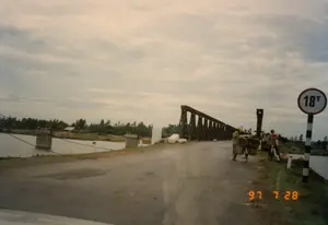 Bức ảnh chụp một con đường nhựa ở vùng nông thôn dẫn đến một cây cầu sắt bắc qua sông. Bầu trời u ám, nhiều mây. Ở bên phải đường, có một biển báo giao thông hình tròn với viền đỏ, nền trắng, ghi chữ '18T', cho biết giới hạn trọng tải của cầu là 18 tấn. Cột của biển báo được sơn sọc đen trắng. Gần biển báo, có hai người dân đang dừng xe đạp. Một người đội mũ cứng màu vàng đang đứng cạnh chiếc xe đạp thồ, chở hai chiếc sọt lớn chứa đầy hàng hóa. Người còn lại mặc áo sơ mi kẻ sọc đỏ, cũng đứng cạnh một chiếc xe đạp. Hoạt động của họ dường như là đang vận chuyển hàng hóa và chuẩn bị qua cầu. Cây cầu là một cây cầu giàn bằng kim loại, có vẻ là một cây cầu tạm thời hoặc cầu phao. Dưới sông, có thể nhìn thấy những trụ cầu bê tông cũ, có lẽ là tàn tích của một cây cầu đã bị phá hủy trước đó. Phía xa bên kia sông là cây cối và một ngôi nhà nhỏ. Góc dưới bên phải của bức ảnh có dấu ngày tháng màu cam '97 7 28'.。（日本語訳）。田舎のアスファルトの道が、川にかかる鉄橋へと続いている写真です。空はどんよりと曇っています。道の右側には、赤い縁取りに白地の円形交通標識があり、「18T」と書かれています。これは橋の重量制限が18トンであることを示しています。標識の柱は白黒の縞模様に塗られています。標識の近くで、2人の地元住民が自転車を止めています。黄色いヘルメットをかぶった一人は、荷物でいっぱいの大きなかごを2つ積んだ荷物運搬用の自転車のそばに立っています。もう一人は赤い縞模様のシャツを着て、同じく自転車のそばに立っています。彼らの様子は、荷物を運んで橋を渡る準備をしているように見えます。橋は金属製のトラス橋で、仮設橋または浮き橋のようです。川の中には、古いコンクリートの橋脚が見えます。おそらく以前に破壊された橋の残骸でしょう。川の向こうの遠くには、木々と小さな家があります。写真の右下隅には、「97 7 28」というオレンジ色の日付スタンプがあります。