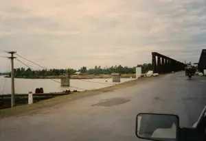 Bức ảnh chụp khung cảnh một con đường nông thôn dẫn lên một cây cầu giàn thép bắc qua sông, có khả năng ở khu vực Quảng Bình hoặc Quảng Trị. Dưới sông có các trụ cầu bê tông cũ, cho thấy cây cầu hiện tại có thể là cầu tạm được dựng lên. Một người đội mũ cối đang đạp xe trên cầu, chở theo hai chiếc giỏ lớn và cồng kềnh ở phía sau. Bên trái đường là một cột điện bằng gỗ và các cọc tiêu màu trắng. Phía xa, bên kia sông, có thể thấy cây cối, một ngôi nhà và một cây rơm. Bức ảnh được chụp từ một chiếc ô tô đang di chuyển, với gương chiếu hậu của xe ở góc dưới bên phải. Không có văn bản nào có thể đọc được.。（日本語訳）。写真は、川に架かる鋼鉄のトラス橋へと続く田舎道の風景を撮影したもので、おそらくクアンビン省またはクアンチ省の地域にあると思われます。川の中には古いコンクリートの橋脚があり、現在の橋が仮設で架けられたものである可能性を示しています。ヘルメットをかぶった人が橋の上を自転車で走っており、後ろに大きくてかさばる籠を2つ運んでいます。道路の左側には、木製の電柱と白いデリニエーターがあります。遠くの川の向こう側には、木々、一軒の家、そして稲わらの山が見えます。読み取れるテキストはありません。