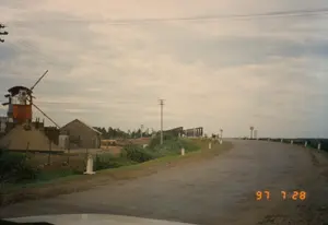 Bức ảnh ghi lại quang cảnh một con đường nhựa ở vùng nông thôn vào ngày 28 tháng 7 năm 1997, được chụp từ bên trong một chiếc xe hơi. Con đường uốn cong về bên phải, hai bên có cắm các cọc tiêu màu trắng. Bên trái đường là một công trường xây dựng hoặc một nhà máy sản xuất vật liệu, với một đống cát/sỏi lớn, một cấu trúc giống si-lô màu cam và một máy móc có cần trục. Cạnh đó là một ngôi nhà cấp bốn. Dọc đường có nhiều cột điện và dây điện. Ở phía xa là một cây cầu có kết cấu giàn thép, có vẻ đang trong quá trình thi công hoặc sửa chữa. Có một vài biển báo giao thông hình tròn ở xa nhưng không thể đọc được nội dung. Bầu trời u ám và nhiều mây. Dấu ngày tháng "97 7 28" được hiển thị ở góc dưới bên phải.。（日本語訳）。1997年7月28日に車内から撮影された、田舎のアスファルト道路の光景を捉えた写真。道路は右にカーブしており、両側には白いデリニエータが立てられている。道路の左側には建設現場か資材製造工場があり、大きな砂/砂利の山、オレンジ色のサイロのような構造物、クレーン付きの機械がある。その隣には平屋の家がある。道沿いには多くの電柱と電線がある。遠くには鉄骨トラス構造の橋があり、建設中か修理中のようだ。遠くにいくつかの円形の交通標識があるが、内容は読み取れない。空は曇っており、雲が多い。右下隅に「97 7 28」の日付スタンプが表示されている。