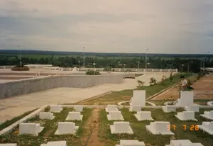 Bức ảnh chụp một không gian rộng lớn, trang nghiêm, dường như là một nghĩa trang liệt sĩ, có thể là một nghĩa trang cấp quốc gia ở khu vực Quảng Bình hoặc Quảng Trị. Ở tiền cảnh, nhiều ngôi mộ liệt sĩ giống hệt nhau được sơn trắng, xếp thành hàng thẳng tắp trên các vạt cỏ. Lối đi giữa các ngôi mộ vẫn còn là đường đất, cho thấy khu vực này có thể đang trong quá trình xây dựng hoặc tu bổ. Ở khu vực trung tâm là một quảng trường lớn lát bê tông với các bức tường và lan can. Một vài cây xanh nhỏ mới được trồng và các cột đèn chiếu sáng được lắp đặt dọc theo lối đi. Có một người đàn ông mặc áo sơ mi trắng đang đứng trong khuôn viên. Phía xa, trải dài đến tận chân trời là khung cảnh nông thôn đặc trưng với những cánh đồng xanh và những lùm cây rậm rạp. Bầu trời u ám, nhiều mây. Không có văn bản nào có thể đọc được trong ảnh.。（日本語訳）。写真は、広大で荘厳な空間を写しており、烈士墓地のようです。おそらくクアンビン省かクアンチ省にある国家レベルの墓地でしょう。前景には、白く塗られた全く同じ形の烈士の墓が多数、芝生の上にまっすぐな列をなして並んでいます。墓の間の通路はまだ土の道であり、この区域が建設中または改修中である可能性を示しています。中央エリアには、壁と手すりのあるコンクリートで舗装された大きな広場があります。いくつかの小さな木が新しく植えられ、通路に沿って照明灯が設置されています。敷地内には白いシャツを着た男性が一人立っています。遠くには、地平線まで広がる、緑の田畑と鬱蒼とした木々が茂る典型的な田園風景が広がっています。空はどんよりとしており、雲が多いです。写真の中には読み取れる文字はありません。