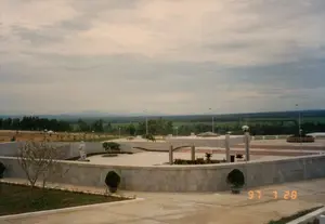 Bức ảnh chụp một khu tưởng niệm hoặc di tích lịch sử rộng lớn ngoài trời, có khả năng là một nghĩa trang liệt sĩ ở khu vực Quảng Bình hoặc Quảng Trị, dựa trên bối cảnh địa lý. Toàn cảnh cho thấy một công trình kiến trúc bằng bê tông và đá với một sân quảng trường lớn ở trung tâm, các bậc tam cấp và một cấu trúc có mái che. Ở phía bên trái của quảng trường có một bức tượng đá trắng. Xung quanh khu vực chính là cây cối, cây cảnh trồng trong chậu và các cột đèn chiếu sáng. Phía sau công trình là một khung cảnh nông thôn rộng lớn với những cánh đồng xanh, cây cối rậm rạp và dãy núi ở phía xa. Bầu trời có nhiều mây. Không có hoạt động nào diễn ra; khung cảnh rất tĩnh lặng và trang nghiêm. Không có văn bản nào có thể đọc được ngoại trừ dấu ngày tháng "97 7 28" ở góc dưới bên phải.。（日本語訳）。この写真は、屋外にある広大な記念碑または史跡を撮影したもので、地理的背景からクアンビン省またはクアンチ省にある烈士墓地である可能性があります。広場の左側には、白い石像があります。主要なエリアの周りには、木々、鉢植えの観葉植物、そして照明灯があります。建物の後ろには、緑の田んぼ、生い茂る木々、そして遠くの山々が広がる広大な田園風景が広がっています。空は曇っています。何の活動も行われておらず、景色は非常に静かで荘厳です。右下隅にある「97 7 28」という日付スタンプを除いて、読み取れる文字はありません。