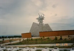 Bức ảnh chụp toàn cảnh một nghĩa trang liệt sĩ quốc gia, rất có thể là Nghĩa trang liệt sĩ quốc gia Trường Sơn ở tỉnh Quảng Trị, vào ngày 28 tháng 7 năm 1997. Ở trung tâm là một Đài Tưởng niệm lớn, hoành tráng được xây bằng đá hoặc bê tông. Trên đỉnh đài tưởng niệm là một cụm tượng điêu khắc trừu tượng hình các chiến sĩ đang trong tư thế chiến đấu. Phía trước đài tưởng niệm và ở tiền cảnh của bức ảnh là khu mộ của các liệt sĩ. Các ngôi mộ được xây bằng xi măng sơn trắng, xếp thành hàng ngay ngắn, trên mỗi mộ có gắn một tấm bia đá màu đen nhưng không thể đọc rõ chữ viết trên đó. Một vài ngôi mộ có cắm hương, cho thấy có người đã đến thăm viếng. Khuôn viên nghĩa trang rộng lớn, có các lối đi và các cây non mới được trồng. Phía sau là những dãy núi và bầu trời u ám, nhiều mây. Dấu ngày tháng '97 7 28' được in ở góc dưới bên phải của bức ảnh.。（日本語訳）。1997年7月28日に撮影された、おそらくクアンチ省にあるチュオンソン国家烈士墓地と思われる、ある国家烈士墓地の全景写真。中央には、石またはコンクリートで造られた大きくて壮大な慰霊塔がある。慰霊塔の頂上には、戦闘態勢にある兵士たちをかたどった抽象的な彫刻群がある。慰霊塔の前、そして写真の前景には、烈士たちの墓地エリアが広がっている。墓は白く塗られたセメントで造られ、整然と列をなしており、それぞれの墓には黒い石の墓碑が取り付けられているが、そこに書かれた文字をはっきりと読み取ることはできない。いくつかの墓には線香が立てられており、誰かが墓参りに来たことを示している。墓地の敷地は広大で、通路や新しく植えられた若木がある。背景には山脈と、暗く曇った空がある。写真の右下隅には「'97 7 28」という日付が印刷されている。