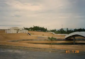 Bức ảnh chụp một cảnh quan rộng lớn dường như là một công trình công cộng hoặc một khu tưởng niệm đang được xây dựng hoặc vừa mới hoàn thành. Dấu thời gian "97 7 28" ở góc dưới bên phải cho thấy ảnh được chụp vào ngày 28 tháng 7 năm 1997. Không có chữ viết nào khác có thể đọc được.。Địa điểm: Đây là một không gian mở, được quy hoạch với địa hình nhân tạo. Mặt đất chủ yếu là đất trống màu nâu đỏ, được san ủi thành nhiều tầng bậc với các bức tường chắn bằng bê tông. Ở phía xa là những hàng cây xanh, cột điện và những dãy núi mờ ảo dưới bầu trời nhiều mây.。Hoạt động: Khung cảnh hoàn toàn tĩnh lặng và không có bóng người, cho thấy công trình chưa được đưa vào sử dụng hoặc đang trong giai đoạn hoàn thiện cảnh quan. Hoạt động chính ở đây là việc xây dựng và tạo dựng cảnh quan quy mô lớn.。Các vật thể: Ở phía bên trái, trên một ngọn đồi thấp, có một công trình kiến trúc hiện đại với mái bằng và các cột trụ màu trắng. Một cây cầu bê tông với thiết kế vòm cong hiện đại bắc qua một con kênh hoặc mương cạn ở phía bên phải. Một vài cây xanh nhỏ mới được trồng, trong đó có một cây ở tiền cảnh được bảo vệ bằng một khung gỗ. Phía trước là một con đường nhựa.。（日本語訳）。この写真は、建設中または完成したばかりの公共事業または記念地区と思われる広大な景観を写しています。右下のタイムスタンプ「97 7 28」は、写真が1997年7月28日に撮影されたことを示しています。他に読み取れる文字はありません。場所：ここは人工的な地形を持つ、計画的に整備されたオープンスペースです。地面は主に赤茶色の空き地で、コンクリートの擁壁で何段にも整地されています。活動：景色は完全に静かで人影はなく、この施設がまだ使用されていないか、景観の仕上げ段階にあることを示しています。ここでの主な活動は、大規模な建設と景観造成です。物体：左側、低い丘の上には、平らな屋根と白い柱を持つ近代的な建築物があります。右側には、近代的なアーチ型のデザインを持つコンクリートの橋が、運河または干上がった水路に架かっています。いくつかの小さな木が新しく植えられており、そのうち前景にある1本は木製のフレームで保護されています。手前にはアスファルトの道路があります。