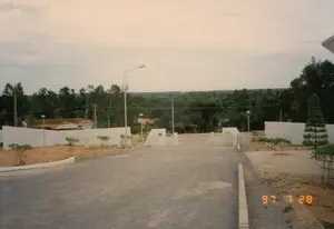 Bức ảnh chụp một con đường rải sỏi dẫn đến một cổng vào lớn được xây bằng bê tông màu trắng. Cánh cổng kim loại đang mở. Hai bên lối vào là tường rào bê tông và khu đất trống đang trong quá trình san lấp, nơi có một số cây non mới được trồng và được chống đỡ bằng khung gỗ. Dọc theo con đường và bên trong khu vực có cổng, có các cột đèn đường. Phía sau bức tường, có thể nhìn thấy một vài ngôi nhà dân với mái ngói đỏ và một lá cờ đỏ (có thể là quốc kỳ Việt Nam) đang bay. Phía xa là khung cảnh vùng nông thôn với những rặng cây xanh và rừng rậm trải dài đến tận chân trời. Bầu trời nhiều mây. Ở góc dưới bên phải ảnh có dấu ngày tháng '97 7 28'.。（日本語訳）。写真には、白いコンクリートで造られた大きな入口の門へと続く、砂利が敷かれた道が写っている。金属製の門は開いている。入口の両側にはコンクリートの塀と、整地中の空き地があり、そこには新しく植えられた何本かの若木が木枠で支えられている。道沿いと門のあるエリアの内側には、街灯の柱がある。遠方には、緑の並木と地平線まで広がる鬱蒼とした森がある田園風景が広がっている。空は曇りが多い。写真の右下隅には「'97 7 28」という日付のスタンプがある。