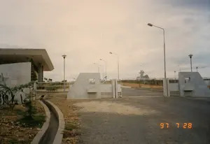 Bức ảnh chụp vào ngày 28 tháng 7 năm 1997, ghi lại quang cảnh lối vào một công trình kiến trúc có vẻ là một khu di tích lịch sử hoặc đài tưởng niệm. Điểm nhấn chính là chiếc cổng lớn, có thiết kế hiện đại, được xây dựng từ các khối bê tông ốp đá màu sáng. Con đường dẫn vào cổng là đường rải sỏi, và hai bên có các cột đèn đường cao. Bên trái có một phần của một tòa nhà cũng mang phong cách kiến trúc hiện đại. Phía sau cổng, trên một ngọn đồi, có một công trình tượng đài lớn với hình dáng độc đáo. Cảnh quan xung quanh còn khá sơ sài với đất và sỏi, cùng một vài cây xanh mới trồng và một rãnh thoát nước bê tông, cho thấy công trình có thể mới được hoàn thành hoặc đang trong giai đoạn hoàn thiện. Bầu trời u ám, không có người hay hoạt động nào được ghi nhận. Không có văn bản hay biển hiệu nào có thể đọc được.。（日本語訳）。1997年7月28日に撮影された写真で、歴史的遺跡または記念碑と思われる建築物への入り口の光景を記録している。主な見どころは大きな門で、モダンなデザインをしており、明るい色の石で覆われたコンクリートブロックで建てられている。門へと続く道は砂利道で、両側には高い街灯が立っている。門の後ろ、丘の上には、ユニークな形状の大きな記念碑がある。周囲の景観は土や砂利でまだかなり簡素で、新しく植えられたいくつかの緑の木々とコンクリートの排水溝があり、この建造物が最近完成したか、あるいは完成段階にあることを示している。空は曇っており、人や活動は記録されていない。読み取れる文字や看板はない。