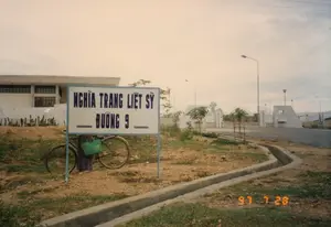 Bức ảnh chụp cảnh quan bên ngoài một nghĩa trang vào một ngày trời u ám. Ở tiền cảnh, một biển báo lớn màu trắng với chữ màu xanh lam được dựng trên bãi đất trống. Trên biển báo có ghi: "NGHĨA TRANG LIỆT SỸ - ĐƯỜNG 9". Dựa vào thông tin này, đây là khu vực Nghĩa trang Liệt sĩ Quốc gia Đường 9. Phía sau biển báo, một chiếc xe đạp cũ được dựng, bên cạnh là một người (dường như là phụ nữ) đang đứng và cầm một chiếc giỏ nhựa màu xanh lá. Một con mương bê tông nhỏ chạy dọc theo rìa của bãi đất. Trong hậu cảnh, có thể thấy một tòa nhà lớn, có thể là nhà tưởng niệm, và xa hơn là cổng vào của nghĩa trang được xây bằng bê tông. Khu vực xung quanh có nhiều cây non mới trồng được chống bằng cọc gỗ và có các cột đèn đường, cho thấy đây là một công trình mới được xây dựng hoặc quy hoạch.。（日本語訳）。曇りの日の、ある墓地の外の風景を撮った写真です。前景には、青い文字が書かれた大きな白い看板が空き地に建てられています。看板には「烈士墓地 - 国道9号線」と書かれています。この情報から、ここは国道9号線国家烈士墓地の区域であることがわかります。看板の後ろには、古い自転車が立てかけられており、その隣には一人の人（女性のようです）が立って、緑色のプラスチック製のカゴを持っています。小さなコンクリートの側溝が空き地の端に沿って走っています。背景には、大きな建物が見えますが、それは記念館かもしれません。そして、さらに遠くにはコンクリートで建てられた墓地の入口の門があります。周辺地域には、木の杭で支えられた新しく植えられた若木が多くあり、街灯の柱もあります。これは、ここが新しく建設または計画された施設であることを示しています。