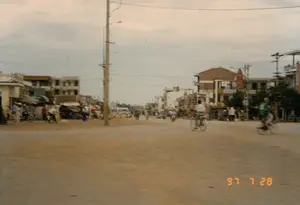 Bối cảnh là một con đường rộng, chưa được trải nhựa tại một thị trấn ở Việt Nam, được chụp vào ngày 28 tháng 7 năm 1997. Con đường bằng đất, có thể là một ngã tư hoặc một quảng trường nhỏ. Hai bên đường là những dãy nhà phố 2-3 tầng mang kiến trúc đặc trưng của thời kỳ đó, một số tòa nhà dường như đang được xây dựng. Nhiều người dân đang sinh hoạt và đi lại trên đường. Phương tiện di chuyển chủ yếu là xe đạp và xe máy. Có thể thấy rõ một người đàn ông mặc áo trắng và một người khác mặc áo xanh đang đạp xe. Ở phía xa có một chiếc xe khách hoặc xe buýt màu trắng. Bên lề đường bên trái, một nhóm người đang tụ tập, có thể là một khu chợ cóc hoặc nơi người dân nghỉ chân. Hệ thống cột điện và dây điện chằng chịt là một đặc điểm nổi bật của khung cảnh đô thị. Có các biển hiệu trên các tòa nhà nhưng không thể đọc rõ nội dung. Dấu ngày tháng "97 7 28" được in ở góc dưới bên phải của hình ảnh.。（日本語訳）。背景は、1997年7月28日に撮影された、ベトナムのある町の未舗装の広い道です。道は土でできており、交差点か小さな広場かもしれません。道の両側には、その時代に特徴的な建築様式の2〜3階建てのタウンハウスが並んでおり、いくつかの建物は建設中のようです。多くの人々が道の上で生活し、行き来しています。主な交通手段は自転車とバイクです。白いシャツを着た男性と、青いシャツを着たもう一人の人が自転車を漕いでいるのがはっきりと見えます。遠くには、一台の白色の長距離バスまたは路線バスがあります。左側の道端では、一団の人々が集まっており、青空市場か、人々が休憩する場所かもしれません。複雑に絡み合った電柱と電線は、この都市景観の際立った特徴です。建物には看板がありますが、その内容ははっきりと読み取れません。画像の右下の角に「97 7 28」という日付が印字されています。