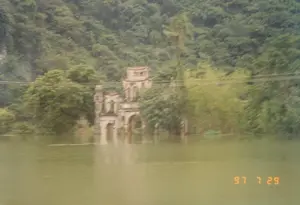 Bức ảnh được chụp vào ngày 29 tháng 7 năm 1997, ghi lại cảnh một công trình kiến trúc cổ, có thể là một nhà thờ hoặc một biệt thự, bị ngập sâu trong nước lũ. Nước lũ có màu xanh đục, dâng cao nhấn chìm phần lớn tầng dưới của tòa nhà. Bối cảnh là một vùng nông thôn với những ngọn đồi hoặc núi được bao phủ bởi cây cối rậm rạp ở phía sau. Một số cây cối, bao gồm một cây dừa, cũng bị ngập một phần trong nước. Có những đường dây điện chạy ngang qua khung hình. Cảnh tượng cho thấy một trận lụt lớn đang diễn ra, không có hoạt động nào của con người được nhìn thấy.。（日本語訳）。写真は1997年7月29日に撮影されたもので、古い建築物、おそらくは教会か別荘が、洪水で深く水に浸かっている光景を記録しています。洪水は濁った緑色で、水位が上がり建物の下層階の大部分を水没させています。背景は田園地帯で、後方には木々が鬱蒼と茂る丘または山があります。ヤシの木を含むいくつかの木々も、部分的に水に浸かっています。電線がフレームを横切っています。この光景は大規模な洪水が発生していることを示しており、人間の活動は見られません。