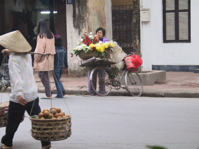 Bức ảnh ghi lại một khung cảnh đường phố sinh động ở Việt Nam. Ở tiền cảnh, một người phụ nữ đội nón lá đang đi bộ trên đường, sử dụng đòn gánh để gánh hai chiếc rổ tre đan. Một trong hai chiếc rổ chứa đầy những quả tròn màu cam, có thể là quất hoặc cam, một mặt hàng phổ biến trong dịp Tết. Ở phía sau, trên vỉa hè, một người phụ nữ khác, có lẽ là người bán hoa, đang đứng cạnh chiếc xe đạp cũ. Chiếc xe đạp được chất đầy các loại hoa tươi nhiều màu sắc như hoa cúc và hoa hồng. Trên ghi đông xe có treo một chiếc giỏ và một cái xô nhựa màu đỏ. Một vài người đi bộ khác cũng xuất hiện trên vỉa hè. Bối cảnh là một dãy nhà có vẻ cũ, với một biển số nhà ghi rõ số '92'. Khung cảnh gợi lên không khí buôn bán tấp nập và sự chuẩn bị cho một ngày lễ, rất có thể là Tết Nguyên Đán.。（日本語訳）。写真はベトナムの活気ある街角の風景を捉えています。前景には、ノンラーをかぶった女性が道を歩いており、天秤棒を使って編んだ竹かごを二つ担いでいます。二つのかごのうちの一つは、オレンジ色の丸い果物でいっぱいです。それはキンカンかオレンジかもしれません。テト（旧正月）の時期によく見られる品物です。後方、歩道の上では、別の女性、おそらく花売りが、古い自転車の隣に立っています。自転車には、菊やバラのような色とりどりの生花がたくさん積まれています。自転車のハンドルには、かご一つと赤いプラスチックのバケツが一つ掛けられています。他にも数人の歩行者が歩道に見られます。背景は古そうな家並みで、「92」という番地がはっきりと書かれたプレートがあります。この光景は、賑やかな商売の雰囲気と祝日の準備を思わせます。おそらくテト（旧正月）でしょう。