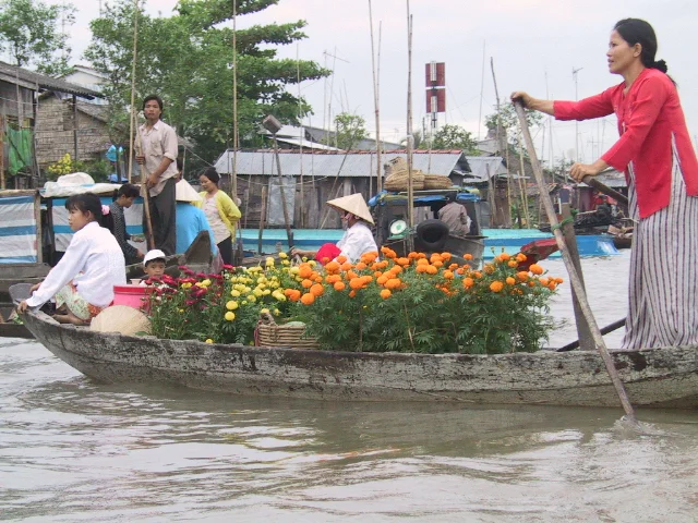 Bối cảnh là một khu chợ nổi trên sông, có thể là ở Cần Thơ, vào dịp gần Tết Nguyên Đán. Một người phụ nữ mặc áo dài cách tân màu đỏ và váy sọc đang đứng chèo một chiếc ghe gỗ. Trên ghe chở rất nhiều chậu hoa cúc vạn thọ màu vàng và cam, cùng một số loại hoa khác, để bán cho người dân mua về chưng Tết. Ngồi ở mũi ghe là một cô gái trẻ mặc áo trắng, và một em bé nhỏ ngồi giữa các chậu hoa. Xung quanh là khung cảnh tấp nập của chợ nổi với nhiều ghe thuyền khác đang qua lại. Ở hậu cảnh, có thể thấy những ngôi nhà ven sông và những người khác, trong đó có người đang đội nón lá, cũng đang hoạt động trên thuyền của họ. Không có chữ viết nào có thể đọc được trong hình.。（日本語訳）。背景は川の水上マーケットで、おそらくカントー、旧正月（テト）が近い時期です。赤いモダンなアオザイと縞模様のスカートを着用した一人の女性が、木製の小舟を立って漕いでいます。舟には、人々がテトの飾りとして買って帰るために、たくさんの黄色やオレンジ色のマリーゴールドの鉢植えと、その他いくつかの種類の花が積まれています。舟の舳先には白い服を着た若い女の子が座っており、花の鉢植えの間には小さな子供が座っています。周りは水上マーケットの賑やかな光景で、他の多くの小舟が行き交っています。背景には、川沿いの家々と、ノンラーを被っている人を含む他の人々が、自分たちの舟の上で活動しているのが見えます。画像の中には読み取れる文字はありません。