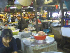 Bối cảnh là một khu chợ trong nhà đông đúc, có thể là một chợ ở Cần Thơ. Không khí rất tấp nập với nhiều người bán hàng và khách mua sắm, gợi đến khung cảnh chuẩn bị cho dịp Tết. Ở phía trước, một người phụ nữ mặc áo sẫm màu đang ngồi bên một bệ hàng tròn, có thể làm bằng xi măng. Trên bệ có một chiếc thau nhựa lớn màu đỏ đựng đầy thực phẩm, trông giống như thịt xay hoặc giò sống. Xung quanh và phía sau là vô số các quầy hàng khác. Các tiểu thương, phần lớn là phụ nữ, đang ngồi sau các sạp hàng của mình, nơi trưng bày nhiều loại thực phẩm trong các thau, rổ, mâm. Trên trần nhà có kết cấu bằng gỗ, có một chiếc nón lá được treo lên. Ánh sáng trong chợ là sự kết hợp giữa ánh sáng tự nhiên và ánh đèn điện. Không có văn bản hay biển hiệu nào có thể đọc được rõ ràng do hình ảnh mờ và lộn xộn.。（日本語訳）。背景は混雑した屋内市場で、おそらくカントーの市場でしょう。多くの売り手と買い物客で大変賑わっており、テト（旧正月）の準備の光景を思わせます。前景には、暗い色の服を着た女性が、おそらくセメントでできた丸い売り台のそばに座っています。周囲と後方には、無数の他の露店があります。小さな商人たち（そのほとんどは女性）は、たらい、かご、盆に様々な食材を陳列した自分の屋台の後ろに座っています。木製の構造をした天井には、ノンラー（ベトナムの伝統的な笠）が一つ吊るされています。市場の照明は、自然光と電灯の光が混ざり合ったものです。画像がぼやけていて雑然としているため、はっきりと読み取れる文字や看板はありません。