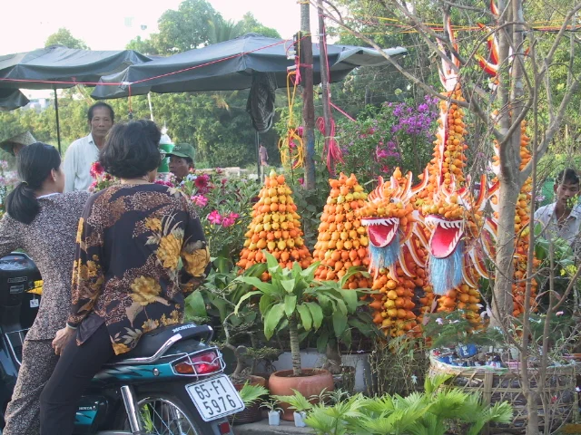 Bối cảnh là một khu chợ hoa Tết ngoài trời, có thể ở Cần Thơ, trong không khí chuẩn bị cho Tết Nguyên Đán. Mọi người đang tham quan và mua sắm các mặt hàng trang trí.。Ở phía trước, hai người phụ nữ đang ngồi trên một chiếc xe máy màu xanh. Người phụ nữ ngồi sau mặc áo hoa văn màu đen và vàng, đang nhìn về phía các quầy hàng.。Điểm nhấn của khung cảnh là các sản phẩm trang trí Tết độc đáo. Có những mô hình con rồng lớn được kết công phu từ rất nhiều quả nhỏ màu cam, có thể là quả cau. Đầu rồng được tạo hình sống động với miệng há rộng. Bên cạnh các con rồng là những tháp trái cây hình chóp cũng được làm từ loại quả tương tự.。Xung quanh còn có rất nhiều chậu cây cảnh và hoa đang được bày bán, như hoa màu hồng và các loại cây xanh khác. Phía sau có một người đàn ông mặc áo sơ mi trắng và một người đàn ông khác đội mũ xanh, có vẻ là người bán hàng hoặc khách mua hàng. Một chiếc dù lớn màu đen che mát cho một phần của khu chợ.。（日本語訳）。背景は、おそらくカントーにある屋外のテト（旧正月）の花市場で、旧正月を迎える準備の雰囲気に包まれています。人々は装飾品を見て回り、買い物をしています。手前では、二人の女性が青いバイクに座っています。後ろに座っている女性は黒と黄色の柄の服を着て、露店の方を見ています。この光景のハイライトは、ユニークなテトの装飾品です。おそらくビンロウの実であろう、たくさんのオレンジ色の小さな果物から精巧に作られた大きな龍の模型があります。龍の頭は生き生きと形作られ、口を大きく開けています。龍の隣には、同じ種類の果物から作られた円錐形の果物の塔があります。周りには、ピンク色の花やその他の緑の植物など、多くの鉢植えの観葉植物や花も販売されています。後ろには、白いシャツを着た男性と青い帽子をかぶった別の男性がおり、店員か買い物客のようです。大きな黒い傘が市場の一角に日陰を作っています。