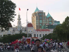 Bối cảnh là một quảng trường hoặc đường phố đông đúc ở trung tâm Thành phố Hồ Chí Minh vào lúc chiều tối. Ở hậu cảnh, có thể thấy rõ tòa nhà trụ sở Ủy ban Nhân dân Thành phố Hồ Chí Minh, một công trình kiến trúc kiểu Pháp với tháp đồng hồ. Trên đỉnh tháp có treo lá cờ đỏ sao vàng của Việt Nam. Phía sau tòa nhà này là một tòa nhà cao tầng hiện đại. Có rất đông người dân đang tụ tập, nhiều người trong số họ đang đi xe máy. Ở giữa đám đông, có những người bán hàng rong đang bán rất nhiều chùm bong bóng bay với đủ màu sắc. Đặc biệt, có những quả bóng bay màu đỏ in biểu tượng búa liềm. Ở phía trước, một người đàn ông đang lái xe máy và chở theo một em bé. Khung cảnh cho thấy một không khí nhộn nhịp, có thể là một dịp lễ hoặc một buổi tối cuối tuần.。（日本語訳）。背景は、夕暮れ時のホーチミン市中心部にある混雑した広場または通りです。背景には、時計塔のあるフランス様式の建築物であるホーチミン市人民委員会庁舎がはっきりと見えます。塔の頂上には、ベトナムの金星紅旗が掲げられています。この建物の後ろには、近代的な高層ビルがあります。多くの人々が集まっており、その多くはバイクに乗っています。人混みの中には、色とりどりの風船の束をたくさん売っている露天商がいます。特に、鎌と槌のシンボルが印刷された赤い風船があります。手前では、一人の男性がバイクを運転し、子供を一人乗せています。その光景は、祝日か週末の夜かもしれない賑やかな雰囲気を示しています。