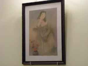 Đây là hình ảnh chụp một bức tranh chân dung được đóng khung và treo trên một bức tường màu kem. Khung tranh có màu đen và được trang trí hoa văn. Bức tranh mô tả một người phụ nữ có mái tóc sẫm màu, đang ngồi và chống cằm vào tay với vẻ mặt trầm tư. Ở góc dưới bên trái của bức tranh có một bình hoa. Bức tranh được thể hiện bằng các tông màu nhẹ nhàng, mờ ảo. Không có bất kỳ văn bản nào có thể đọc được trong hình ảnh.。（日本語訳）。これは、額装され、クリーム色の壁に掛けられた肖像画の写真です。額縁は黒色で、模様で装飾されています。この絵画は、暗い色の髪をした女性が座り、物思いにふける表情で手に顎を乗せている様子を描いています。絵画の左下隅には花瓶があります。この絵画は、柔らかく、ぼんやりとした色調で表現されています。画像内には読み取れるテキストはありません。