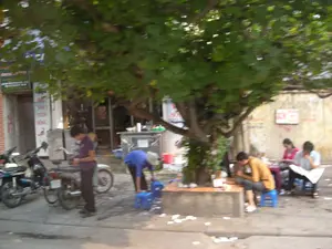 Bức ảnh ghi lại một cảnh sinh hoạt trên vỉa hè ở Việt Nam, dưới một gốc cây lớn. Đây có vẻ là một quán ăn vỉa hè.。Về chữ viết, có một biển hiệu dọc màu trắng với chữ đỏ, có thể đọc được một phần là "... SẢN TƯƠI SỐNG". Ngoài ra còn có các biển hiệu khác ở phía bên trái và trên bức tường phía sau nhưng bị mờ và không thể đọc được nội dung.。Về khung cảnh và hoạt động, một nhóm người đang ngồi trên những chiếc ghế nhựa màu xanh lam quanh một chiếc bàn thấp để ăn uống. Một người đàn ông đang ăn, một người khác thì đang đọc báo. Một người mặc áo xanh đang cúi người làm việc, có thể là chuẩn bị đồ ăn hoặc dọn dẹp gần một quầy hàng bằng kim loại. Nhiều chiếc xe máy đang được đỗ trên vỉa hè. Khung cảnh cho thấy một hoạt động buôn bán ăn uống đường phố rất đặc trưng.。（日本語訳）。この写真は、ベトナムの歩道で、大きな木の下での日常の光景を捉えています。これは歩道の屋台のようです。文字については、赤文字の白い縦長の看板があり、「... SẢN TƯƠI SỐNG」と一部読み取れます。その他にも、左側や後ろの壁に他の看板がありますが、ぼやけていて内容は読み取れません。風景と活動については、数人のグループが低いテーブルを囲んで青いプラスチックの椅子に座り、食事をしています。ある男性は食事をしており、別の人は新聞を読んでいます。青いシャツを着た人が、金属製の屋台の近くで、おそらく料理の準備か片付けのために、かがんで作業をしています。歩道には多くのバイクが駐車されています。この光景は、非常に特徴的な路上での飲食販売の活動を示しています。