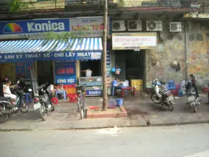 Bức ảnh chụp một cảnh đường phố ở Việt Nam với các cửa hàng san sát nhau. Bên trái là một cửa hàng ảnh có biển hiệu lớn màu xanh ghi chữ 'Konica', 'FILM & CAMERAS' và 'Ảnh Màu NẮNG XUÂN'. Phía dưới mái hiên sọc xanh trắng có một biển hiệu khác ghi 'CHỤP ẢNH KỸ THUẬT SỐ - CHỜ LẤY NGAY'. Bên cạnh, một cửa hàng khác có biển hiệu màu trắng ghi 'CHUYÊN BÁN BUÔN BÁN LẺ THỦY HẢI SẢN' và số điện thoại '0912256196'. Trên vỉa hè, nhiều xe máy đang được đậu. Một bé gái mặc đồ trắng đang ngồi trên yên một chiếc xe máy. Gần đó, một người đàn ông đang ngồi ăn trên chiếc ghế nhựa thấp. Một chiếc xe đạp được dựng cạnh gốc cây ở giữa vỉa hè. Phía bên phải, một người phụ nữ đang đứng cạnh một chiếc xe máy. Trên bức tường cũ của tòa nhà bên phải có treo một chiếc nón lá và lắp đặt một vài cục nóng điều hòa.。（日本語訳）。この写真はベトナムのある通りの風景を写したもので、店が隣接して並んでいます。左側には写真店があり、「Konica」、「FILM & CAMERAS」、「Ảnh Màu NẮNG XUÂN」と書かれた大きな青い看板があります。青と白のストライプの庇の下には、「CHỤP ẢNH KỸ THUẬT SỐ - CHỜ LẤY NGAY」と書かれた別の看板があります。その隣には、別の店があり、「CHUYÊN BÁN BUÔN BÁN LẺ THỦY HẢI SẢN」と電話番号「0912256196」が書かれた白い看板があります。歩道には、多くのバイクが停められています。白い服を着た女の子がバイクのサドルに座っています。その近くでは、男性が低いプラスチックの椅子に座って食事をしています。歩道の中央にある木の根元には、自転車が立てかけられています。右側では、女性がバイクの隣に立っています。右側の建物の古い壁には、ノンラーが一つ掛けられ、エアコンの室外機がいくつか設置されています。