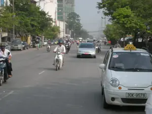 Cảnh tượng trên một con đường phố đông đúc tại Việt Nam. Dòng xe cộ tấp nập với nhiều loại phương tiện, chủ yếu là xe máy và ô tô. Mọi người đang điều khiển phương tiện của mình để tham gia giao thông, và hầu hết những người đi xe máy đều đội mũ bảo hiểm. Ở phía trước bên phải, có một chiếc xe taxi màu trắng. Trên nóc xe có biển hiệu màu vàng ghi chữ 'TAXI'. Trên kính chắn gió của xe taxi có một miếng dán hình tròn màu đỏ ghi số '130'. Dọc hai bên đường là những hàng cây xanh và các tòa nhà san sát. Ở phía xa có thể thấy một tòa nhà cao tầng đang được xây dựng. Có một số biển hiệu trên các tòa nhà bên đường nhưng không thể đọc rõ nội dung.。（日本語訳）。ベトナムの混雑した通りの光景。車両の流れはひっきりなしで、主にバイクや自動車など様々な種類の乗り物がある。人々は交通に参加するために自分の乗り物を運転しており、バイクに乗る人のほとんどはヘルメットを着用している。右手前には、白いタクシーが一台ある。車の屋根には「TAXI」と書かれた黄色い看板がある。タクシーのフロントガラスには、「130」という数字が書かれた赤い円形のステッカーがある。道の両脇には緑の並木と建物がびっしりと建ち並んでいる。道沿いの建物にはいくつかの看板があるが、その内容をはっきりと読み取ることはできない。