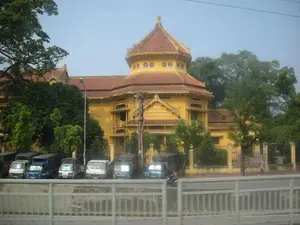 Bức ảnh ghi lại khung cảnh một tòa nhà lớn, có kiến trúc cổ điển, sơn màu vàng đặc trưng và lợp mái ngói đỏ. Tòa nhà này mang đậm phong cách kiến trúc Đông Dương, nổi bật với phần tháp trung tâm hình bát giác. Xung quanh tòa nhà được bao bọc bởi nhiều cây xanh. Phía trước, trong khuôn viên được rào lại, có một dãy xe tải nhỏ đang đỗ thành hàng. Bên ngoài là một con đường có xe máy đang lưu thông và một người đi bộ trên vỉa hè. Một lan can bê tông nằm ở tiền cảnh của bức ảnh. Không có chữ viết hay biển hiệu nào có thể nhận dạng được.。（日本語訳）。写真には、古典的な建築様式を持ち、特徴的な黄色に塗られ、赤い瓦屋根で葺かれた大きな建物の光景が写っている。この建物はインドシナ建築様式を色濃く反映しており、八角形の中央の塔が際立っている。建物の周りは多くの緑の木々に囲まれている。前方の、柵で囲まれた敷地内には、小さなトラックが一列に駐車している。外側にはバイクが通行している道路と、歩道を歩く一人の歩行者がいる。写真の前景にはコンクリートの手すりがある。認識できる文字や看板はない。