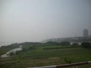 Bức ảnh chụp một khung cảnh rộng lớn nhìn từ trên cao, có thể là từ một cây cầu, bao quát một khu vực ven sông. Bầu trời có nhiều sương mù, làm cho cảnh vật ở xa, bao gồm các tòa nhà của một thành phố, trở nên mờ ảo. Ở phía trước và trung tâm là những cánh đồng nông nghiệp xanh tươi, được chia thành nhiều thửa ruộng để trồng trọt. Bên trái là một con sông lớn hoặc một hồ nước. Dọc theo bờ sông ở phía xa là một khu đô thị với nhiều nhà cửa và một vài tòa nhà cao tầng. Khung cảnh cho thấy sự giao thoa giữa vùng nông thôn với đất canh tác và khu vực đô thị đang phát triển. Không có chữ viết nào có thể nhận dạng được trong ảnh.。（日本語訳）。この写真は、おそらく橋の上から撮影された、川沿いの地域を見渡す広大な風景を捉えています。空は霧が多く、遠くにある街の建物を含む景色がぼやけて見えます。手前と中央には、耕作のために多くの区画に分けられた、青々とした農地が広がっています。左側には大きな川または湖があります。遠くの川岸に沿って、多くの家屋といくつかの高層ビルがある都市部があります。この風景は、農地のある田園地帯と発展途上の都市部との交わりを示しています。写真には識別できる文字はありません。