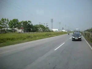 Trên nóc chiếc xe taxi màu đen có biển hiệu ghi chữ 'TAXI' và 'MAI LINH'. Phía sau, trên một chiếc xe khách màu xanh dương có chữ, có thể là tên nhà xe như 'HẢI ÂU'.。Đây là khung cảnh trên một con đường quốc lộ hoặc đường lớn ở khu vực ngoại ô. Một chiếc xe taxi màu đen đang di chuyển trên đường, theo sau là một chiếc xe khách màu xanh và một người đi xe máy. Bên lề đường bên trái là một khu đất trống có cỏ mọc và nhiều cây xanh. Phía sau hàng cây là một vài công trình nhà ở hoặc nhà xưởng có hàng rào. Dọc con đường có các cột điện và đường dây điện cao thế. Ở phía xa có thể nhìn thấy một chiếc xe trộn bê tông và các công trình khác. Bầu trời u ám.。（日本語訳）。黒いタクシーの屋根の上には、「TAXI」と「MAI LINH」と書かれた看板があります。後方には、青いバスに文字が書かれており、それは「HẢI ÂU」のようなバス会社の名前かもしれません。これは郊外の国道または大きな道路の光景です。黒いタクシーが道路を走行しており、その後ろには青いバスとバイクに乗った人が続いています。道路の左側には、草が生え、多くの緑の木々がある空き地があります。木々の列の後ろには、フェンスのある住宅または工場の建物がいくつかあります。道路沿いには電柱と高圧電線があります。遠くには、コンクリートミキサー車やその他の建造物が見えます。空は曇っています。