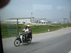 Bức ảnh chụp cảnh trên một con đường nhựa ở một khu vực có vẻ là ngoại ô hoặc gần khu công nghiệp. Ở tiền cảnh, một người đàn ông và một người phụ nữ đang đi trên một chiếc xe máy. Người đàn ông đang lái xe, mặc áo sơ mi trắng, đội mũ bảo hiểm màu tối và đeo kính râm. Người phụ nữ ngồi sau, mặc áo hoa văn và đội mũ bảo hiểm màu vàng. Chiếc xe máy có một giỏ ở phía trước. Bên cạnh con đường là một khu đất trồng cỏ xanh. Ở phía sau, có một khu nhà xưởng hoặc nhà máy lớn, hiện đại với tường trắng và hàng rào màu xanh. Một lá cờ Việt Nam đang bay trên một trong các tòa nhà. Có các cột điện và đường dây điện chạy dọc theo khu vực. Ở phía xa, có một người dường như đang làm việc trên cánh đồng. Không có văn bản nào có thể đọc được trên các tòa nhà.。（日本語訳）。写真は、郊外または工業団地の近くと思われる地域の、アスファルトの道路上の風景を撮影したものです。前景には、男性と女性がバイクに乗っています。男性が運転しており、白いシャツを着て、暗い色のヘルメットをかぶり、サングラスをかけています。女性は後ろに乗っており、柄物の服を着て、黄色のヘルメットをかぶっています。バイクの前部にはかごが付いています。道路の脇には、緑の草が生えた土地があります。後方には、白い壁と緑のフェンスがある、大きくて近代的な工場またはプラントがあります。建物の1つにベトナムの国旗がはためいています。この地域に沿って電柱と電線が走っています。建物には読み取れる文字はありません。