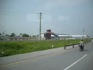 Bức ảnh chụp quang cảnh bên ngoài một khu công nghiệp từ một con đường nhựa. Trên cổng chào lớn có kiến trúc hình vòm màu đỏ và trắng, có dòng chữ "KHU CÔNG NGHIỆP ĐẠI AN". Bên trong hàng rào của khu công nghiệp là các tòa nhà nhà xưởng, một tháp nước có treo lá cờ Việt Nam và một số cây cọ. Dọc theo con đường là một bãi cỏ và lan can bảo vệ. Một người đàn ông đang lái xe máy trên đường. Phía trên là bầu trời nhiều mây cùng với hệ thống cột điện và dây điện.。（日本語訳）。アスファルトの道路からある工業団地の外の光景を撮影した写真。大きな歓迎ゲートには赤と白のアーチ型の建築があり、「KHU CÔNG NGHIỆP ĐẠI AN」という文字が書かれている。工業団地のフェンスの内側には、工場の建物、ベトナム国旗が掲げられた給水塔、そして数本のヤシの木がある。道路に沿って芝生のエリアとガードレールがある。一人の男性が道路でバイクを運転している。上方には、電柱と電線のシステムと共に雲の多い空が広がっている。