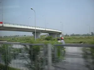Bức ảnh ghi lại khung cảnh một cây cầu vượt bằng bê tông dành cho đường bộ. Bức ảnh được chụp từ một phương tiện đang di chuyển, thể hiện qua phần tiền cảnh bị mờ, bao gồm một dải phân cách và mặt đường ướt. Trên con đường song song, có hai người đang đi xe máy, một người đội mũ bảo hiểm màu đỏ. Dưới gầm cầu, trên một bãi cỏ rộng lớn, có một vài người đang ngồi nghỉ. Dọc theo cây cầu và con đường là các cột đèn đường. Ở phía xa là những cánh đồng và một vài công trình xây dựng, nhà cửa.。（日本語訳）。写真は、道路用のコンクリート製高架橋の光景を記録したものです。写真は移動中の乗り物から撮影されたもので、中央分離帯と濡れた路面を含むぼやけた前景にそれが表れています。並行する道では、二人がバイクに乗っており、一人は赤いヘルメットをかぶっています。橋の下の広い芝生の上では、数人が座って休んでいます。橋と道に沿って街灯があります。遠くには田畑といくつかの建設物、家屋があります。