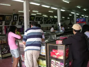 Bên trong một cửa hàng lớn, có thể là một cửa hàng lưu niệm hoặc trạm dừng chân. Không gian rộng, có nhiều cột trụ, trần nhà lắp nhiều đèn tuýp và quạt trần. Nhiều người, có vẻ là khách du lịch, đang xem và lựa chọn hàng hóa được trưng bày trong các tủ kính. Một người đàn ông mặc áo thun sọc và một phụ nữ mặc áo hồng đang đứng xem các sản phẩm. Một người khác đội mũ màu be cũng đang xem hàng ở quầy bên cạnh. Trên tường treo rất nhiều tranh ảnh nghệ thuật được đóng khung. Các tủ kính trưng bày nhiều loại hàng hóa, bao gồm bánh kẹo và các bức tượng nhỏ. Có thể đọc được chữ 'HÒA AN' và 'Choco Chips' trên các hộp sản phẩm. Ở phía sau có một tủ lạnh màu đỏ để bán nước giải khát.。（日本語訳）。大きな店の中、それは土産物店か休憩所かもしれない。多くの人々、見たところ観光客のようだが、ガラスケースに陳列された商品を見て選んでいる。ストライプのTシャツを着た男性とピンクの服を着た女性が立って製品を見ている。ベージュ色の帽子をかぶった別の人物も隣のカウンターで商品を見ている。壁には額装された芸術的な絵画や写真がたくさん掛けられている。ガラスケースには、菓子や小さな像など、多くの種類の商品が陳列されている。商品の箱には「HÒA AN」や「Choco Chips」という文字が読み取れる。後方には飲料を販売するための赤い冷蔵庫がある。