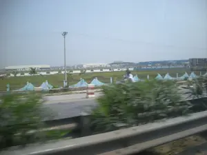 Bức ảnh được chụp từ một phương tiện đang di chuyển, nhìn ra khung cảnh ven đường. Ở phía trước, các bụi cây và lan can bảo vệ bị mờ do tốc độ di chuyển. Có một con đường nhựa, trên đó một người đang đi xe máy. Dọc theo con đường là một hàng rào trang trí màu xanh nhạt và một cọc tiêu giao thông sọc đỏ trắng. Phía sau hàng rào là một khu đất rộng lớn có cỏ và một vài cây cọ nhỏ mới trồng. Ở phía xa là các tòa nhà lớn, có thể là nhà máy hoặc một phần của khu công nghiệp, cùng với một cột điện và đường dây điện. Bầu trời quang đãng. Không có văn bản nào có thể đọc được trong ảnh.。（日本語訳）。写真は移動中の乗り物から撮影されたもので、道端の風景を眺めている。手前では、茂みとガードレールが移動速度のためにぼやけている。アスファルトの道路があり、その上をバイクに乗った人が一人走っている。道路に沿って、水色の装飾的なフェンスと、赤と白の縞模様の交通ポールがある。フェンスの後ろには、草が生えた広大な土地と、新しく植えられた数本の小さなヤシの木がある。空は晴れている。写真には読み取れる文字はない。