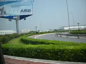 Bức ảnh được chụp từ một phương tiện đang di chuyển, nhìn ra một con đường nhựa uốn lượn trong một khu công nghiệp hoặc khu đô thị mới. Cảnh quan hai bên đường được quy hoạch gọn gàng với các hàng rào cây xanh được cắt tỉa, bãi cỏ và các bụi hoa. Ở phía bên trái, có một biển quảng cáo lớn với nội dung: "AMB và Hải Phòng bắt tay để xây dựng một Thành phố Quốc tế." cùng với logo "AMB" và tên công ty "amco-mibaek vina co.,ltd.". Ở phía xa, có thể thấy các tòa nhà lớn, hiện đại, có khả năng là nhà máy hoặc tòa nhà văn phòng. Một trong những tòa nhà này có logo "Pioneer" ở mặt tiền. Dọc theo con đường có các cột đèn đường kiểu cách. Bầu trời có vẻ hơi u ám.。（日本語訳）。写真は移動中の乗り物から撮影されたもので、工業団地か新興都市区の中にある、曲がりくねったアスファルトの道が写っている。道の両側の景観はきれいに整備されており、刈り込まれた生け垣、芝生、そして花壇がある。左側には、「AMBとハイフォンが手を取り合い、国際都市を建設する。」という内容の大きな広告看板があり、「AMB」のロゴと「amco-mibaek vina co.,ltd.」という会社名も添えられている。遠くには、大きくて近代的な建物が見え、それらは工場かオフィスビルである可能性がある。これらの建物のうちの一つには、正面に「Pioneer」のロゴがある。空は少し曇っているようだ。