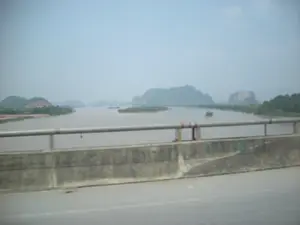 Bức ảnh được chụp từ trên một cây cầu, nhìn ra khung cảnh một con sông rộng lớn. Dòng sông có màu nước đục phù sa. Ở giữa sông có một cù lao nhỏ và có một vài chiếc thuyền, tàu đang di chuyển, có thể là tàu vận chuyển hàng hóa. Phía xa là phong cảnh núi non trập trùng. Phần tiền cảnh của bức ảnh là lan can bê tông của cây cầu, trên đó có vẽ những con số màu xanh lá cây nhưng không thể đọc rõ, và một con đường nhựa.。（日本語訳）。写真は橋の上から撮影されたもので、広大な川の風景が見えます。川の水は沖積土で濁っています。川の中ほどには小さな中州があり、数隻の船やボートが航行していて、それらは貨物船かもしれません。遠くには、幾重にも連なる山々の風景が広がっています。写真の前景は、緑色の数字が描かれているものの、はっきりと読み取れない橋のコンクリートの欄干と、アスファルトの道路です。