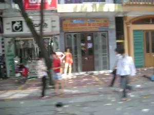 Bức ảnh bị mờ, chụp cảnh một con phố với các cửa hàng liền kề. Có một số biển hiệu có thể đọc được một phần. Một biển hiệu màu vàng ở giữa ghi rõ tên 'NGỌC TÚN' và mặt hàng kinh doanh là 'TỔ YẾN - VI CÁ CAO CẤP'. Phía trên bên trái, một biển hiệu màu đỏ có chữ 'WINY'. Một cửa hàng khác trưng bày hai ma-nơ-canh ở phía trước, một ma-nơ-canh mặc váy đỏ, gợi ý đây là một cửa hàng thời trang. Trên vỉa hè có một vài người đang đi lại, nhưng hình ảnh của họ bị nhòe do chuyển động. Một chiếc xe máy màu đỏ đang đậu ở bên trái. Một thân cây lớn đứng ở tiền cảnh, che khuất một phần các cửa hàng. Toàn bộ khung cảnh cho thấy một hoạt động đường phố thường ngày tại một khu vực thương mại.。（日本語訳）。写真はぼやけており、隣接する店舗が並ぶ通りを写している。部分的に読み取れる看板がいくつかある。中央にある黄色い看板には『NGỌC TÚN』という名前と、取扱商品である『高級ツバメの巣 - フカヒレ』がはっきりと書かれている。左上には、『WINY』という文字が入った赤い看板がある。別の店は正面に2体のマネキンを展示しており、1体は赤いドレスを着ていて、ここがファッション店であることを示唆している。歩道には数人の人が歩いているが、その姿は動きによってぼやけている。左側には赤いバイクが1台停まっている。前景には大きな木の幹が立っており、店舗の一部を覆い隠している。全体の光景は、商業地域における日常的な通りの活動を示している。