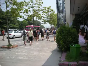 Bối cảnh là một bãi đỗ xe vào một ngày nắng. Một nhóm người, có vẻ là khách du lịch nước ngoài, đang đi bộ qua bãi đỗ xe. Họ mặc trang phục mùa hè như quần short, áo phông và mang ba lô. Nhóm người này đang đi về phía một chiếc xe buýt du lịch màu hồng đang đỗ ở phía sau. Trong bãi đỗ xe còn có các phương tiện khác như một chiếc ô tô con màu trắng và nhiều xe khác ở phía xa. Bên phải là một tòa nhà có mặt tiền bằng kính lớn. Có một số người đang đứng và ngồi gần lối vào tòa nhà. Phía trước tòa nhà có một bụi cây và một thùng rác màu xanh lá cây. Cây xanh được trồng dọc theo bãi đỗ xe.。（日本語訳）。背景は晴れた日の駐車場です。外国人観光客と思われる一団が、駐車場を歩いて渡っています。彼らはショートパンツやTシャツといった夏服を着て、バックパックを背負っています。この一団は、後方に駐車しているピンク色の観光バスの方へ向かっています。駐車場には、白い乗用車や遠くにある他の多くの車など、他の車両もあります。右側には、大きなガラス張りのファサードを持つ建物があります。建物の入り口の近くで、何人かの人が立ったり座ったりしています。建物の前には、茂みと緑色のゴミ箱があります。駐車場に沿って緑の木々が植えられています。