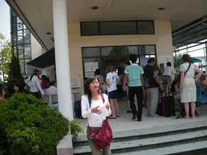 Bối cảnh là một bến tàu du lịch, có khả năng là ở khu vực Vịnh Hạ Long. Phía trước là một tòa nhà có mái hiên rộng, nơi một nhóm đông người, bao gồm cả du khách Việt Nam và người nước ngoài, đang tụ tập.。**Chữ viết:**。Trên cửa kính ra vào có dòng chữ lớn màu đỏ: 'KHÁCH LIÊN HỆ HỢP ĐỒNG' và bên dưới là 'TÀU DU LỊCH'. Bên cạnh cửa có dán một số giấy tờ, thông báo nhưng không thể đọc rõ nội dung.。**Cảnh vật và Hoạt động:**。Địa điểm này là một nhà ga hoặc bến tàu dùng để phục vụ khách du lịch đi thuyền. Mọi người đang đứng và ngồi chờ đợi, có vẻ như đang chuẩn bị cho một chuyến đi thuyền hoặc vừa kết thúc chuyến đi. Một số người đang trò chuyện với nhau. Có một chiếc vali màu đen đỏ đặt trên sàn, cho thấy đây là nhóm khách du lịch. Một người phụ nữ mặc áo sơ mi trắng, đeo túi xách màu đỏ ở tiền cảnh dường như là một hướng dẫn viên du lịch đang nói chuyện hoặc hướng dẫn cho nhóm của mình. Phía sau, có những dãy ghế chờ bằng nhựa màu xanh. Xung quanh có cây cảnh được trồng để trang trí. Mọi người đều mặc trang phục mùa hè, thoải mái.。（日本語訳）。背景は観光船の船着き場で、おそらくハロン湾エリアにあると思われます。前方には広い庇のある建物があり、そこにはベトナム人観光客と外国人を含む大勢のグループが集まっています。**文字：**。出入り口のガラス戸には、「契約に関するご連絡」という大きな赤い文字があり、その下には「観光船」と書かれています。ドアの横にはいくつかの書類や通知が貼られていますが、内容ははっきりと読み取れません。**風景と活動：**。この場所は、船旅をする観光客にサービスを提供するためのターミナルまたは船着き場です。人々は立ったり座ったりして待っており、船旅の準備をしているか、あるいは旅を終えたばかりのようです。何人かの人々は互いに会話しています。床には赤と黒のスーツケースが一つ置かれており、これが観光客のグループであることを示しています。前景にいる、白いシャツを着て赤いハンドバッグを提げた女性はツアーガイドのようで、自分のグループに話しかけたり、案内したりしています。後方には、青いプラスチック製の待合椅子が並んでいます。周りには装飾のために観葉植物が植えられています。誰もが快適な夏服を着ています。