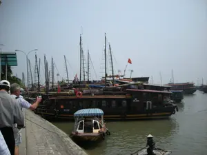 Quang cảnh tại một bến tàu du lịch, có thể là ở khu vực Vịnh Hạ Long. Dọc theo cầu tàu bằng đá là rất nhiều tàu du lịch lớn bằng gỗ, được thiết kế theo kiểu thuyền buồm truyền thống với nhiều cột buồm, đang neo đậu san sát nhau. Trên thân các con tàu có thể thấy các số hiệu như "QN-2988" và "QN-1988". Một con tàu có chữ "TOURIST" ở mạn thuyền. Một lá cờ Việt Nam được treo trên một trong những con tàu.。Dưới mặt nước, có một chiếc xuồng máy nhỏ có mái che sọc xanh trắng đang cập sát vào một tàu lớn. Ở phía trước, một người đội nón lá đang chèo một chiếc thuyền nan nhỏ.。Trên cầu tàu, một nhóm khách du lịch đang đứng quan sát, trong đó có một người đàn ông đang dùng máy ảnh để chụp lại khung cảnh.。（日本語訳）。ある観光船の船着き場の光景で、おそらくハロン湾エリアのものでしょう。石造りの桟橋に沿って、多くのマストを持つ伝統的な帆船の様式で設計された、たくさんの木製大型観光船が密集して停泊しています。船体には「QN-2988」や「QN-1988」のような船体番号が見えます。一隻の船の船腹には「TOURIST」という文字があります。船の一隻にはベトナム国旗が掲げられています。水面では、青と白の縞模様の屋根が付いた小型モーターボートが、大型船に接岸しています。手前では、ノンラーをかぶった人が小さな竹の船を漕いでいます。桟橋の上では、観光客のグループが立って見物しており、その中にはカメラで景色を撮影している男性が一人います。